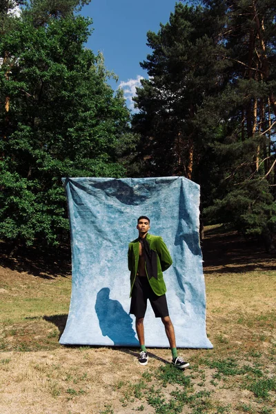 African american man in green blazer and black shorts standing with hands behind back near blue drape in park — Photo de stock