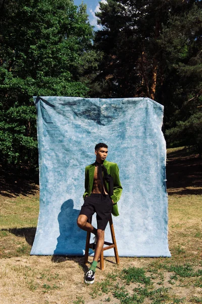 Full length of african american man in green blazer sitting with hands in pockets near blue cloth in forest — стоковое фото