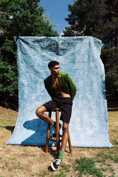 African american man in green blazer and back shorts posing on stool near blue drape in forest — Stock Photo