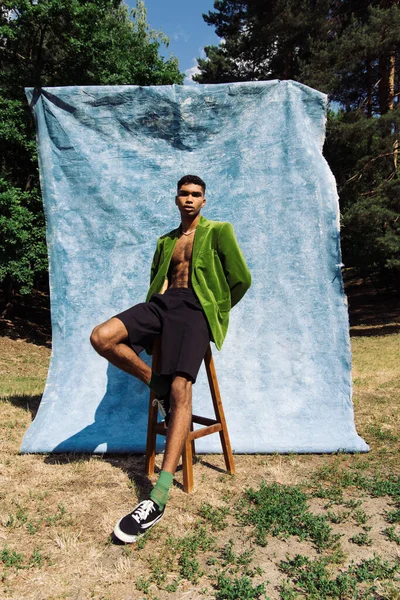 Модний афроамериканський чоловік в оксамитовому блістері і кросівки, сидячи на табуреті біля синьої тканини в парку — стокове фото