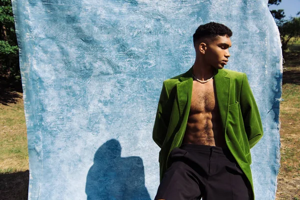 African american man in green velvet blazer sitting with hands behind back near blue cloth — Foto stock