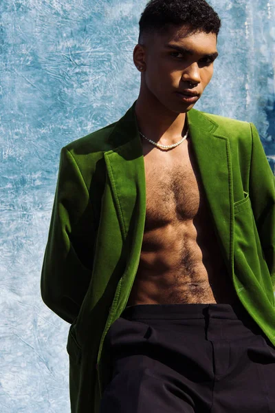 Young african american man in green blazer and beads looking at camera near blue cloth on background — Fotografia de Stock