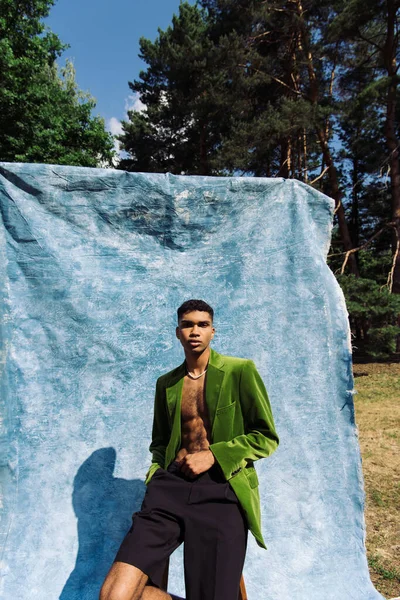 African american man in velvet blazer sitting with hand in pocket near blue drape in park — стоковое фото