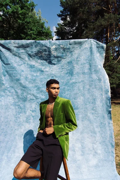 African american man in green blazer and black shorts sitting with closed eyes in park near blue cloth - foto de stock