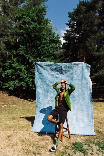 African american man in trendy clothes sitting on stool with hands on head in park near blue drapery — стоковое фото