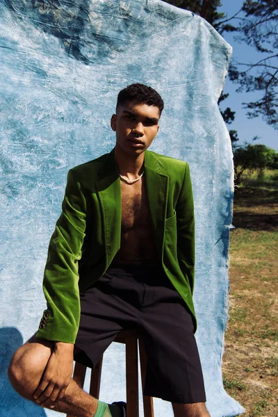 African american man in black shorts and green blazer sitting in park near blue drape — Stock Photo