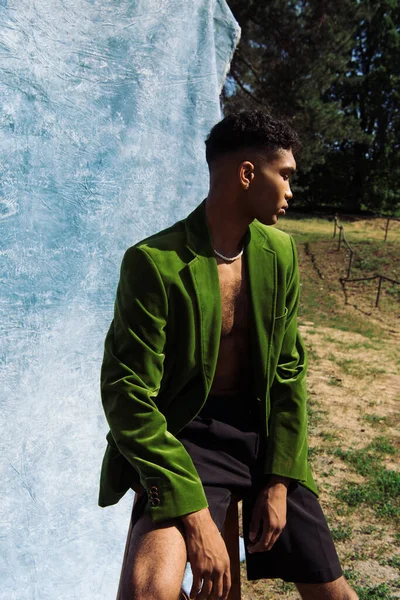 Young african american man in green velvet blazer and black shorts sitting in forest near blue drapery — Stockfoto