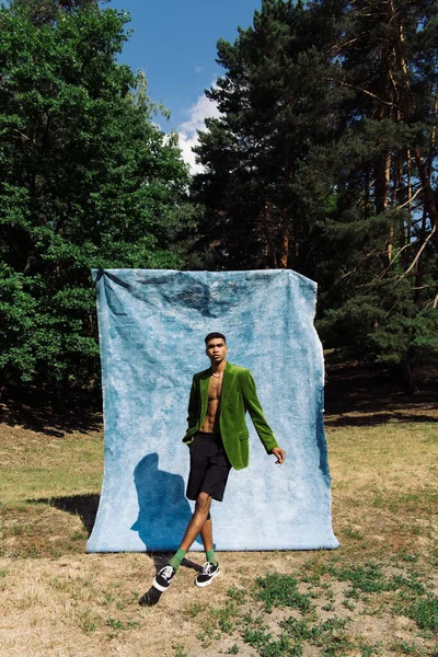 Full length of african american man in green blazer and sneakers standing with hand in pocket near blue cloth in park — Stock Photo