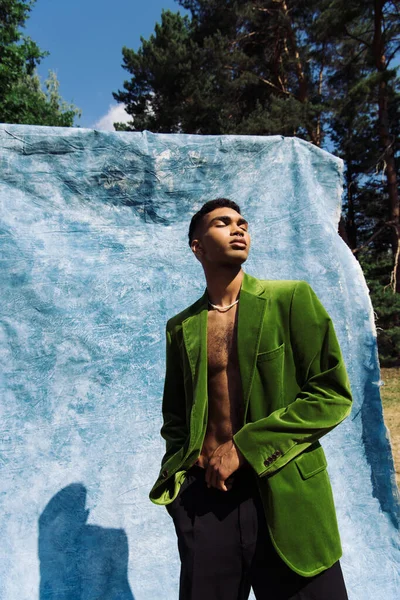 African american man in green velvet blazer posing with closed eyes near blue cloth — Stockfoto