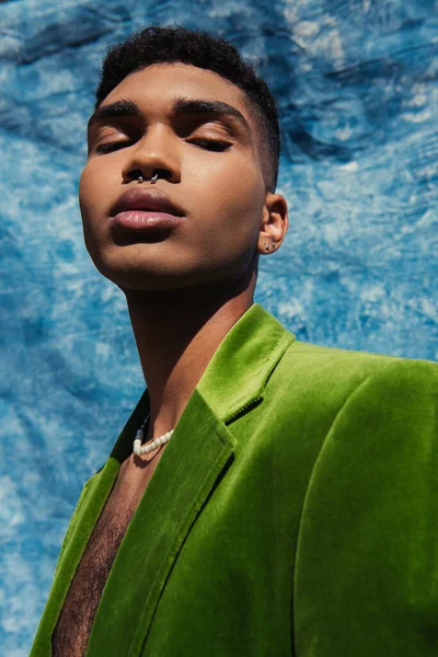 African american man with piercing posing in green blazer and beads near blue drape — стоковое фото