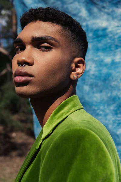 Portrait of african american man with piercing looking at camera near blue cloth on blurred background — Stockfoto