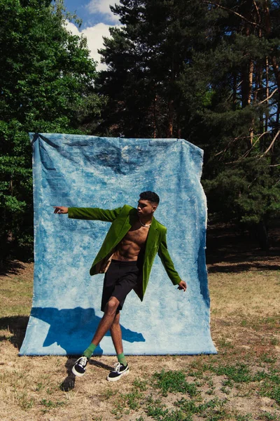 Trendy african american man in velvet blazer and shorts pointing with finger near blue drape in forest — Fotografia de Stock