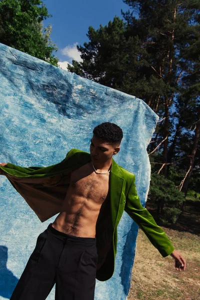 Trendy african american man in green velvet blazer posing in park near blue drapery — Stockfoto