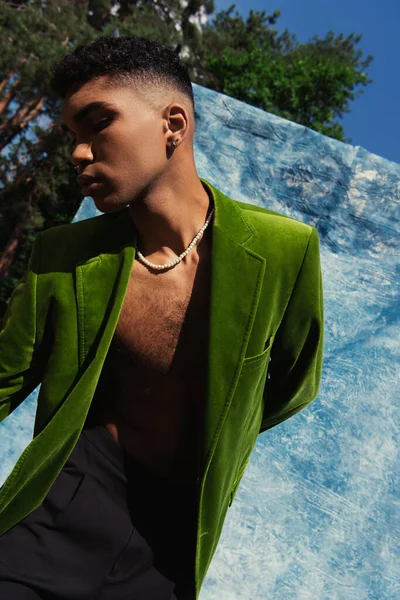 Fashionable african american man posing in green blazer near blue cloth in park - foto de stock