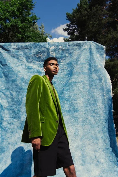 African american man in green stylish blazer looking at camera near blue drapery in park - foto de stock