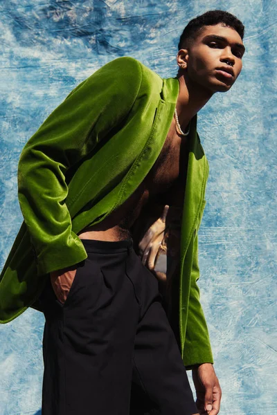 Low angle view of african american man in green velvet blazer posing near blue drapery on background — Foto stock