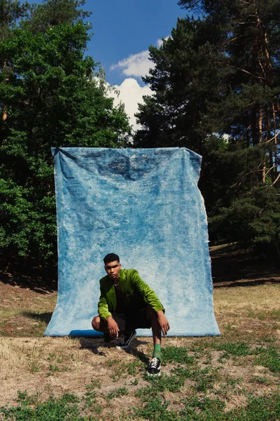 Fashionable african american man sitting on haunches in forest near blue drapery — Stock Photo