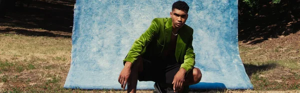 Young african american man in green velvet blazer posing near blue textured cloth outdoors, banner — Stockfoto