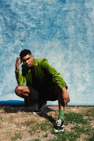 Full length of african american man in green blazer and sneakers posing on haunches near blue drapery — Stock Photo