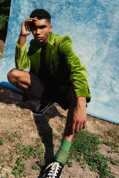 African american man in green blazer and sneakers looking at camera near blue cloth outdoors — стоковое фото