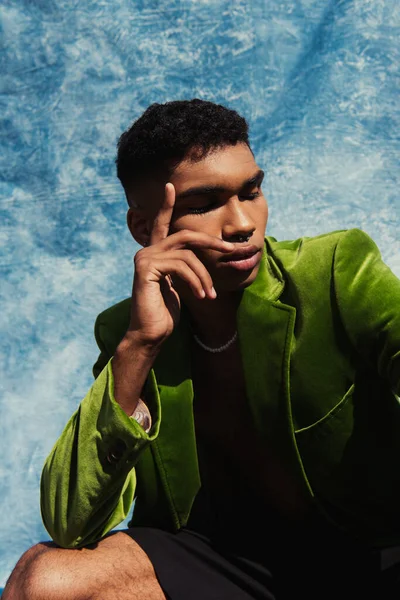 Young african american man in green blazer holding hand near face while posing with closed eyes near blue cloth — Fotografia de Stock