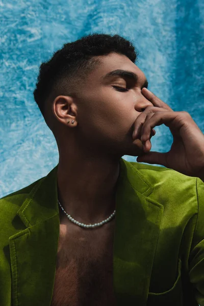 Stylish african american man in green blazer and beads covering face with hand near blue drape on background — стоковое фото