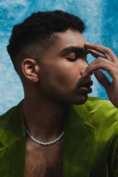 Young african american man in green velvet blazer and beads obscuring face near blue drapery — Foto stock