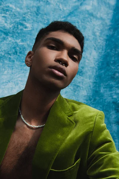 Trendy african american man with piercing and beads look at camera near blue drapery — Stockfoto