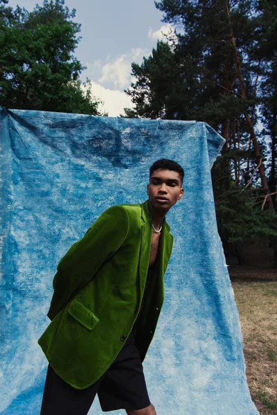 Young african american man in green velvet blazer standing with hand behind back near blue drape in forest — Stock Photo