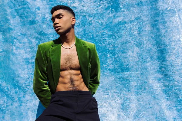 Young african american man in fashionable green blazer posing with hands behind back near blue drape - foto de stock