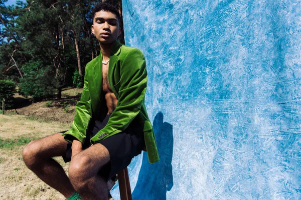 Stylish african american man in green velvet blazer sitting with closed eyes near blue drape in forest - foto de stock