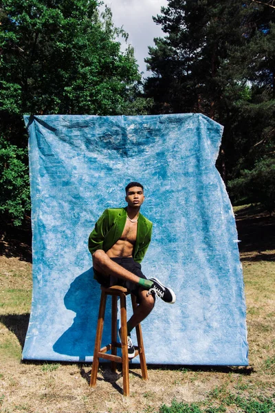 Full length of african american man in blazer and sneakers sitting on stool in park near blue drapery — Photo de stock