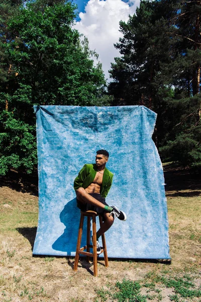 Full length of african american man in black shorts and green blazer posing on stool in forest near blue cloth — стоковое фото