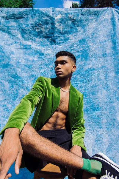 Fashionable african american man looking at camera while sitting near blue drape outdoors — Photo de stock