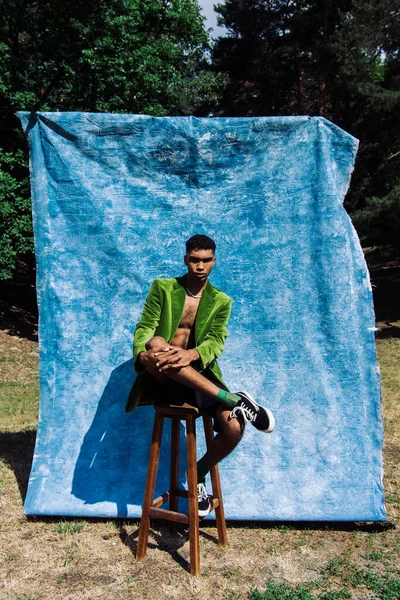 Full length of african american man in green blazer and sneakers sitting in park near blue drapery - foto de stock