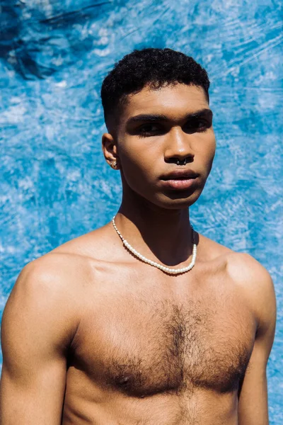 Shirtless african american man with piercing and beads looking at camera near blue drape — Fotografia de Stock