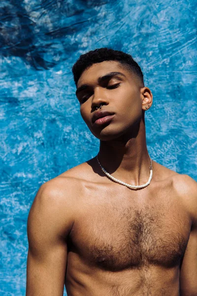 Shirtless african american man in beads and piercing posing with closed eyes near blue cloth — Photo de stock