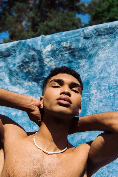 Low angle view of young african american man posing with hands behind neck near blue drapery — Stockfoto