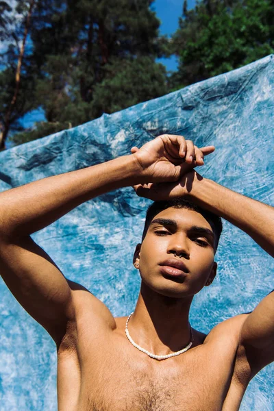 Low angle view of shirtless african american in beads posing near blue cloth outdoors — Photo de stock