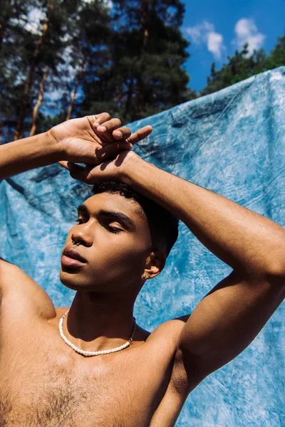 Молодий афроамериканський чоловік з голими грудьми і пірсинг позує руками над головою біля синьої тканини — стокове фото