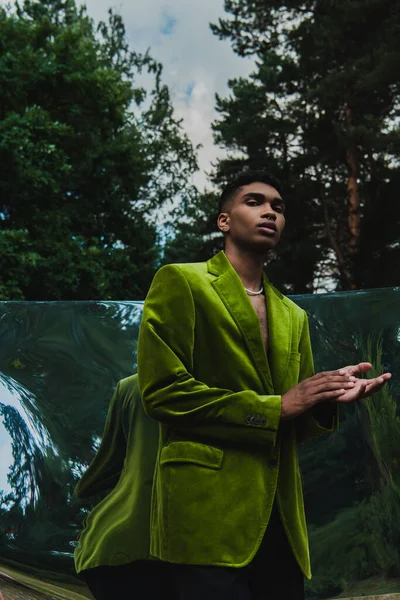 Low angle view of african american man in green velvet blazer looking at camera near reflective surface in park - foto de stock