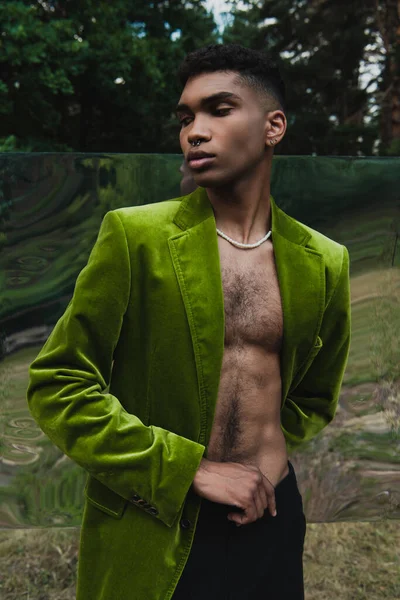 Fashionable african american man in beads and velvet blazer posing in forest near flexible mirror — Stock Photo