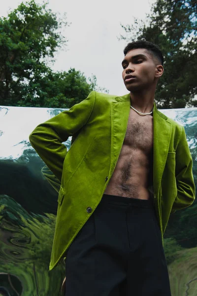 Trendy african american man in green blazer standing with hands behind back near mirror surface in forest — стоковое фото