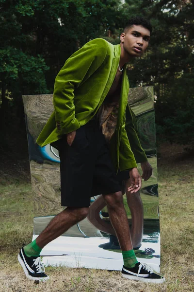 Full length of african american man in green blazer and hand in pocket of black short walking near mirror in park — стоковое фото