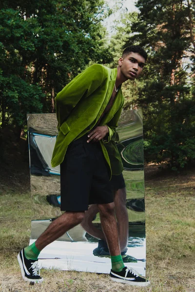 Full length of african american man in black shorts and green velvet blazer walking near flexible mirror in park — Stockfoto