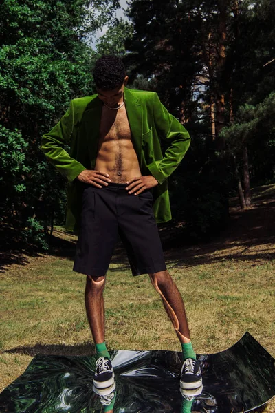 Full length of african american man in green blazer and black shorts standing on mirror surface in park - foto de stock