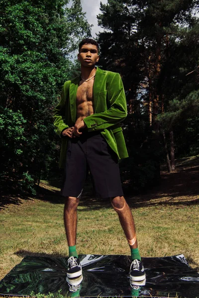 Full length of african american man in black shorts and green blazer standing on specular surface in park — Photo de stock