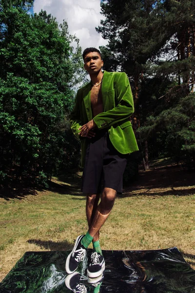 Full length of african american man in blazer and sneakers standing on mirror surface in forest - foto de stock