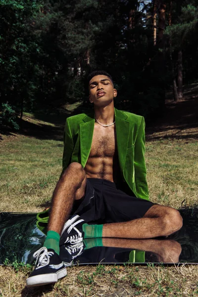 Trendy african american man in velvet blazer and sneakers sitting on mirror in park - foto de stock