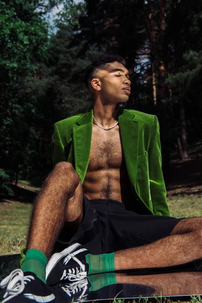 Young and stylish african american man in green blazer and black shorts sitting on mirror surface in forest - foto de stock